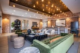 Located at 5060 City Station Dr in Collegedale, Tennessee, this single-floor community amenity space blends industrial and contemporary design into a cohesive, inviting environment. The open-plan layout is anchored by rich light hardwood flooring that flows throughout, unifying the various distinct zones within the space.

The lounge area features a tufted velvet sofa and curated seating arranged around a sleek linear fireplace, with a wall-mounted flat-screen TV above. French doors on either side flood the area with natural light and provide direct access to an outdoor patio. A striking coffered ceiling with warm wood paneling and cascading Edison bulb pendant lights adds architectural character and visual depth.

The dining and social zone showcases exposed brick accent walls that lend an industrial warmth to the space. A live-edge wood dining table paired with contemporary chairs serves as a focal point, complemented by a long white quartz bar with barstool seating. Additional pendant lighting in matte black with gold interiors illuminates a generous banquette upholstered in caramel leather against teal-painted walls.

Throughout the space, decorative brick columns, arched shelving displays, and curated artwork contribute to a layered, upscale aesthetic. The thoughtful combination of textures, materials, and lighting creates a versatile gathering space equally suited for casual socializing or structured group use.