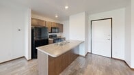 This single-floor residence offers a practical and comfortable layout across three bedrooms, two bathrooms, a kitchen, and a dedicated laundry room. The open-concept living and kitchen area serves as the heart of the home, featuring wood-look hard flooring, a ceiling fan, and a sliding glass door that leads to an outdoor balcony. The kitchen is equipped with a peninsula island featuring a double-basin sink with a laminate countertop, dark wood cabinetry, and black appliances including a refrigerator, range, and over-the-range microwave. The living area transitions to plush carpeting and is filled with natural light. Each of the three bedrooms is carpeted and features a window with open views of the surrounding landscape and neighborhood. The primary bedroom offers a comfortable footprint with recessed lighting. Both bathrooms are finished with wood-toned tile flooring, dark wood vanities with granite-style countertops, and full soaking tub and shower combinations with white surrounds. The dedicated laundry room houses a full-size top-loading washer and dryer. Throughout the space, white walls, warm wood trim accents, and recessed lighting create a clean, cohesive aesthetic.