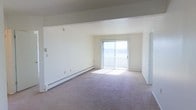 This single-floor space offers a clean, functional layout with a focus on simplicity and natural light. The main living area is generously sized, featuring neutral wall-to-wall carpeting that contributes to a warm, understated atmosphere. Crisp white walls keep the space feeling open and bright throughout.

A prominent sliding glass door serves as a central architectural feature of the living area, fitted with vertical blinds and allowing abundant natural light to fill the room. The door provides direct access to an outdoor area, enhancing the sense of openness and connection to the exterior.

Baseboard heating runs along the lower perimeter of the main room, ensuring efficient climate control throughout the space. Structural beams are visible near the ceiling, adding a subtle architectural detail to the otherwise clean-lined interior. A secondary door is also present, offering additional access or transition to another area of the space.

Overall, the space presents a practical and well-lit environment. The neutral color palette, open layout, and natural light create a versatile foundation well-suited to a variety of uses and furnishing styles.