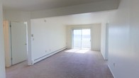 This single-floor space offers a clean, functional layout with a focus on simplicity and natural light. The main living area is generously sized, featuring neutral wall-to-wall carpeting that contributes to a warm, understated atmosphere. Crisp white walls keep the space feeling open and bright throughout.

A prominent sliding glass door serves as a central architectural feature of the living area, fitted with vertical blinds and allowing abundant natural light to fill the room. The door provides direct access to an outdoor area, enhancing the sense of openness and connection to the exterior.

Baseboard heating runs along the lower perimeter of the main room, ensuring efficient climate control throughout the space. Structural beams are visible near the ceiling, adding a subtle architectural detail to the otherwise clean-lined interior. A secondary door is also present, offering additional access or transition to another area of the space.

Overall, the space presents a practical and well-lit environment. The neutral color palette, open layout, and natural light create a versatile foundation well-suited to a variety of uses and furnishing styles.