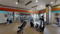 This fitness center is a thoughtfully designed workout space that combines functionality with a vibrant aesthetic. The room is defined by bold horizontal stripe accents in orange and teal that run along the upper walls, creating an energetic atmosphere well-suited for exercise. Recessed lighting and ceiling fans are integrated into coffered ceiling details, ensuring the space remains well-lit and comfortable during workouts.

Large arched windows along one wall admit generous natural light while offering views to the exterior, enhancing the open, airy feel of the room. Full-length mirrors line a significant portion of the walls, visually expanding the space and allowing users to monitor their form during exercise.

The fitness center is comprehensively equipped with a wide variety of machines and accessories. Cardio offerings include elliptical trainers, treadmills, and stationary bikes. Strength training is supported by cable and weight machines, a dedicated dumbbell rack spanning multiple weight increments, adjustable benches, and TRX suspension training straps suspended from the ceiling. A wall-mounted television provides entertainment, and a sanitation station near the entrance promotes cleanliness. The speckled rubberized flooring throughout provides durability and cushioning underfoot.