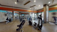 This fitness center is a thoughtfully designed workout space that combines functionality with a vibrant aesthetic. The room is defined by bold horizontal stripe accents in orange and teal that run along the upper walls, creating an energetic atmosphere well-suited for exercise. Recessed lighting and ceiling fans are integrated into coffered ceiling details, ensuring the space remains well-lit and comfortable during workouts.

Large arched windows along one wall admit generous natural light while offering views to the exterior, enhancing the open, airy feel of the room. Full-length mirrors line a significant portion of the walls, visually expanding the space and allowing users to monitor their form during exercise.

The fitness center is comprehensively equipped with a wide variety of machines and accessories. Cardio offerings include elliptical trainers, treadmills, and stationary bikes. Strength training is supported by cable and weight machines, a dedicated dumbbell rack spanning multiple weight increments, adjustable benches, and TRX suspension training straps suspended from the ceiling. A wall-mounted television provides entertainment, and a sanitation station near the entrance promotes cleanliness. The speckled rubberized flooring throughout provides durability and cushioning underfoot.