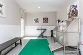 Located at 10 Rimington Lane in Decatur, Georgia, this single-floor professional pet grooming studio is thoughtfully designed to provide a functional and welcoming environment for pets and their owners alike. The space is organized into distinct grooming zones, each outfitted with purpose-built equipment to support a full range of grooming services.

The main grooming area features a stainless steel grooming tub with an integrated ramp for easy pet access, accompanied by a wall-mounted blow dryer for efficient drying. A hydraulic lift grooming table with an adjustable arm provides a dedicated space for styling and finishing work. The flooring is smooth and easy to clean, with a green anti-fatigue mat providing comfort underfoot. A door with glass panels connects the grooming zones, allowing natural light to flow between areas.

The reception or waiting area is finished with warm neutral tones and classic board-and-batten wainscoting, giving it a polished, welcoming feel. A simple wooden bench offers seating, and a glass-panel exterior door allows natural light to brighten the space.

Throughout the studio, vibrant and colorful dog-themed artwork — including bold pop-art portraits of bulldogs and pugs — adds personality and charm to the otherwise clean, professional interior. Recessed lighting and ceiling vents ensure a comfortable environment. Overall, the studio balances practicality with a fun, pet-friendly atmosphere.