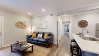 Located at Pointe Grand in Athens, GA, this single-floor residence offers a thoughtfully designed open-concept layout finished with warm wood-look flooring, crown molding, and a neutral color palette throughout. The living area provides a welcoming atmosphere, anchored by recessed lighting, decorative wall accents, and a cozy seating arrangement that flows naturally into the rest of the unit. A well-lit hallway connects the living spaces to the private bedrooms, each of which features ample natural light from large windows, soft neutral tones, and direct access to a bathroom. The bathrooms are outfitted with crisp white cabinetry, wood-framed mirrors, and clean, bright finishes. The kitchen is a standout feature, showcasing white shaker-style cabinetry, a bold blue subway tile backsplash, stainless steel appliances, and a central island with a built-in sink and bar seating — ideal for both cooking and casual dining. A dedicated dining nook with a custom wall graphic adds a personalized touch to the space. Crown molding and recessed lighting are consistent throughout, lending a polished, cohesive aesthetic. The overall ambiance is modern yet warm, combining contemporary design elements with comfortable, functional living.