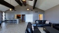 This single-story space makes a strong architectural statement with its soaring vaulted ceilings, accented by bold, dark-stained exposed wood beams that draw the eye upward and add striking visual contrast against the light gray walls. A ceiling fan is centrally mounted, providing both function and proportion to the expansive vertical space above.

The open-concept layout seamlessly connects the living area to the kitchen, promoting a sense of spaciousness and ease of flow between the two zones. The living area is anchored by dark leather sofas arranged to encourage conversation, complemented by light tile flooring that extends throughout the space. The neutral gray wall color creates a clean, cohesive backdrop.

The kitchen features warm wood cabinetry, a tiled breakfast bar with seating, and dark appliances, offering a functional and inviting cooking space that remains visually connected to the main living area. A set of double closet doors is positioned along the back wall, providing convenient storage access. An additional doorway leads to further areas of the home. The overall ambiance is open, modern, and casual, with architectural details that give the space a distinctive character.