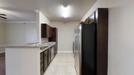 This single-floor space showcases a well-appointed galley-style kitchen as its centerpiece. The kitchen is thoughtfully designed with rich, dark espresso-toned cabinetry that extends from floor to ceiling, providing generous storage along both sides of the corridor layout. Light granite countertops offer a striking contrast against the dark wood tones, while a stainless steel sink is neatly integrated into the countertop surface. Stainless steel appliances, including a refrigerator and range, complement the modern aesthetic and add a polished, cohesive look to the space. The light-colored ceramic tile flooring runs the length of the kitchen, lending a clean and bright feel underfoot. Overhead, flush-mounted lighting fixtures illuminate the space effectively, ensuring a well-lit environment for cooking and meal preparation. The neutral, light-toned walls create an open and airy atmosphere that balances the darker cabinetry. The kitchen's linear layout maximizes efficiency, with ample counter space and cabinetry arranged for practical workflow. The textured ceiling adds a subtle traditional touch to the otherwise contemporary design, giving the overall space a comfortable and functional character.