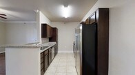 This single-floor space showcases a well-appointed galley-style kitchen as its centerpiece. The kitchen is thoughtfully designed with rich, dark espresso-toned cabinetry that extends from floor to ceiling, providing generous storage along both sides of the corridor layout. Light granite countertops offer a striking contrast against the dark wood tones, while a stainless steel sink is neatly integrated into the countertop surface. Stainless steel appliances, including a refrigerator and range, complement the modern aesthetic and add a polished, cohesive look to the space. The light-colored ceramic tile flooring runs the length of the kitchen, lending a clean and bright feel underfoot. Overhead, flush-mounted lighting fixtures illuminate the space effectively, ensuring a well-lit environment for cooking and meal preparation. The neutral, light-toned walls create an open and airy atmosphere that balances the darker cabinetry. The kitchen's linear layout maximizes efficiency, with ample counter space and cabinetry arranged for practical workflow. The textured ceiling adds a subtle traditional touch to the otherwise contemporary design, giving the overall space a comfortable and functional character.