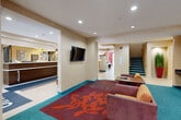 Located in Westborough, MA, this hotel common area offers a well-appointed, generously sized single-floor layout designed to serve guests with comfort and functionality. The front lobby greets visitors with a dark wood reception desk, recessed lighting, large-format tile flooring, and vibrant patterned area rugs in teal, red, and orange that anchor the space with a contemporary energy. A decorative wall clock and colorful artwork add character throughout. The lounge area provides comfortable seating with plush sofas and armchairs arranged around a wall-mounted flat-screen TV, ideal for relaxing or informal gatherings. A secondary seating nook features a continuous light blue upholstered bench along warm-toned walls with teal carpeting. The breakfast and dining area includes a full buffet station with granite-topped counters, built-in warming equipment, and sneeze guards, flowing into a dining room with bold crimson accent walls, arched windows that invite natural light, and a mix of booth and table seating. An ATM alcove with decorative framed artwork provides added convenience. Staircase access and clear circulation paths connect all areas efficiently, while consistent warm beige wall tones and accent lighting unify the overall aesthetic throughout this functional hospitality space.
