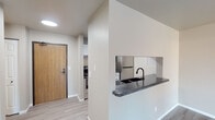 This single-floor unit offers a clean, modern aesthetic throughout, with light gray luxury vinyl plank flooring and neutral warm-toned walls creating a cohesive and inviting atmosphere across all rooms. The open-concept living area is spacious and versatile, bathed in natural light from a sliding glass door and equipped with a wall-mounted air conditioning unit and baseboard heating for year-round comfort. A dedicated bedroom features its own window with a view to the outdoors, baseboard heating, and ample room for a variety of furniture arrangements. The kitchen is thoughtfully designed with white shaker-style cabinetry accented by matte black hardware, dark stone-look countertops, stainless steel appliances including a full-size refrigerator, and a black undermount sink set into a peninsula that opens toward the living area — ideal for entertaining or casual dining. A mirrored stainless steel backsplash adds a sleek, contemporary touch to the kitchen space. The bathroom is well-appointed with a white vanity, dark-framed mirror, combination tub and shower, and coordinating matte black fixtures that tie the space together. A keypad-secured entry door with warm wood tones adds both security and character. Overall, the unit presents a well-maintained, move-in-ready space with consistent modern finishes throughout.