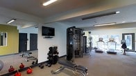 This fitness center is thoughtfully designed to provide a complete exercise environment within a single, open-plan floor. The space features clean white walls and a neutral, rubberized flooring that creates a practical and focused workout atmosphere. Overhead fluorescent lighting ensures the entire area is well-lit throughout the day.

The strength training section is stocked with a tiered dumbbell rack holding a wide range of free weights, a kettlebell, a multi-function cable machine, and a dedicated weight bench — offering versatility for both beginners and experienced users. A wall-mounted flat-screen television is positioned for convenient viewing during workouts.

The cardio zone, situated toward the perimeter of the space, benefits from natural light streaming in through large windows. It is equipped with multiple cardio machines, including a treadmill, an upright exercise bike, and an elliptical, allowing users to choose from a variety of aerobic exercises.

A mirrored wall on one side enhances the sense of space and assists with form and technique during training. Access to the facility is provided through a secure dark-paneled door, adding a clean, modern aesthetic to the overall design. The layout is efficient and well-organized, making the most of the available space.