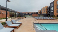 This outdoor amenity courtyard is situated within a contemporary multi-story building complex defined by a striking combination of warm brick and dark metal panel facade materials. The space is designed as an open-air retreat, enclosed on multiple sides by the surrounding structure, creating a sheltered yet sky-exposed environment.

The courtyard centers around a spa and hot tub, with stainless steel entry rails and clear blue water serving as the focal point of the space. Extending outward from the water feature, a spacious concrete deck provides ample room for relaxation and movement. Numerous wood-framed chaise lounges with cushioned blue-grey upholstery are arranged throughout the deck, offering comfortable seating oriented to make the most of the open courtyard setting.

Toward the rear of the space, a lounge seating area with sofas and low tables provides a more social gathering zone, partially shaded by the building's architectural overhangs. Large floor-to-ceiling glass windows and sliding doors line the ground-floor interior spaces adjacent to the courtyard, enhancing the indoor-outdoor connection. Landscape plantings line the building perimeter, softening the hardscape and adding a natural element to the overall design. Overhead lighting poles ensure the space remains functional into the evening hours.