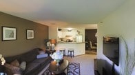 This single-floor residence offers a well-organized, open-concept layout that seamlessly connects the main living areas. The living room is comfortably appointed with plush seating, a decorative area rug, and a flat-screen television, creating a welcoming space for relaxation. Warm, earthy tones on the accent walls complement the neutral carpeted flooring, contributing to a cozy and inviting ambiance throughout.

The kitchen is centrally positioned and features white cabinetry, a refrigerator, a range with an overhead microwave, and a peninsula countertop with bar-height seating — ideal for casual dining or entertaining. The white cabinetry and light countertops keep the kitchen feeling bright and functional.

Adjacent to the kitchen, a dedicated dining area provides additional space for seated meals, furnished with a classic chair and decorative lighting. The open sightlines between the living room, kitchen, and dining area enhance the sense of spaciousness and allow for easy flow between zones.

Natural and artificial lighting work together to keep the interior well-lit. Decorative touches such as floral arrangements, framed wall art, and accent lamps add personality and warmth to the space, making it feel thoughtfully curated and move-in ready.