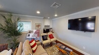 This single-floor space is thoughtfully designed to blend comfort and functionality. The main lounge area serves as the focal point, featuring rich hardwood flooring, a patterned area rug, and recessed lighting set against a gray ceiling accented with crisp white crown molding. A wall-mounted flat-screen television anchors one wall, while a neutral sofa adorned with red, white, and blue accent pillows and a pair of tufted accent chairs provide comfortable seating. A live-edge coffee table adds a natural, organic touch to the arrangement.

A built-in kitchenette area is equipped with white beadboard cabinetry extending to the ceiling, granite countertops with subtle speckled detailing, a stainless steel undermount sink, and a subway tile backsplash. A beverage refrigerator and coffee station make this area well-suited for hosting. Natural light filters through slatted window blinds, illuminating the space warmly.

A dedicated workspace with upper white cabinetry and a desktop workstation is seamlessly integrated into the lounge area. A hallway connects to additional rooms and features a large decorative world map, adding a distinctive visual element. The overall ambiance is polished yet inviting, with a cohesive neutral color palette punctuated by purposeful decorative accents.