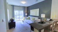 This single-floor residence offers a thoughtfully designed open-concept layout that seamlessly blends living and dining areas. The living room is a standout feature, anchored by a sleek white sofa accented with green throw pillows and complemented by a glass coffee table that adds a light, airy feel to the space. A bold accent wall in deep charcoal gray serves as a striking backdrop, with a large canvas artwork adding a vibrant focal point. Plush carpeting throughout the living area provides warmth and comfort underfoot. Natural light flows generously into the space through expansive floor-to-ceiling sliding glass doors, which open directly onto a private balcony furnished with outdoor seating — ideal for relaxing and enjoying exterior views. Patterned chevron drapes frame the glass doors, adding a dynamic design element. The dining area features a dark-toned table with contemporary styling, creating a natural transition between the living and kitchen spaces. Recessed and flush-mount ceiling lighting ensures the interior remains bright and welcoming. Overall, the space presents a clean, modern aesthetic with a cohesive color palette of neutral tones, grays, and green accents throughout.