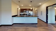 This single-floor unit presents a cohesive, modern design characterized by a neutral color palette, dark trim accents, and a thoughtful mix of flooring materials throughout. The open-concept living and kitchen area forms the heart of the unit, where wood-look plank flooring flows seamlessly beneath a well-appointed kitchen. The kitchen is outfitted with rich, dark cabinetry, dark granite countertops, and a full suite of stainless steel appliances, including a French door refrigerator, range, and microwave. A peninsula with pendant lighting overhead provides both additional prep space and a natural division between the kitchen and living area, which is illuminated by a flush-mount ceiling fixture and benefits from natural light through a window with exterior views.

The carpeted bedrooms offer a quieter, softer environment, each equipped with a ceiling fan and windows that invite natural light. Doors from the bedroom areas connect to adjoining spaces, including a bathroom featuring dark granite countertops and clean, neutral walls. Throughout the unit, dark baseboards and door frames provide a consistent design detail that anchors the lighter wall tones, lending the overall space a polished and contemporary feel.