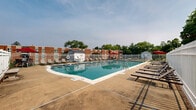 Located at 303 W State St in Doylestown, PA, this outdoor pool area offers a well-maintained recreational facility designed for relaxation and leisure. The centerpiece is a large, rectangular swimming pool filled with clear, blue-green water, accessible via stainless steel entry rails. The pool is surrounded by a wide concrete sundeck that provides ample space for movement and lounging. Numerous lounge chairs are arranged along the perimeter, offering plenty of seating for sunbathing and relaxation. Pops of color are provided by red patio umbrellas, creating shaded spots for those seeking relief from the sun. The entire pool area is enclosed by a white metal fence, ensuring safety and privacy. Pool accessories, including flotation rings and foam noodles, are stored near a poolside utility shed. Signage indicates that swimming is unsupervised, with no lifeguard on duty. The surrounding landscape features lush green lawns and mature trees, providing a pleasant natural backdrop. Views from the pool include neighboring brick residential buildings and a white structure, situating this amenity within a well-established community setting in the heart of Bucks County, Pennsylvania.
