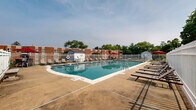 Located at 303 W State St in Doylestown, PA, this outdoor pool area offers a well-maintained recreational facility designed for relaxation and leisure. The centerpiece is a large, rectangular swimming pool filled with clear, blue-green water, accessible via stainless steel entry rails. The pool is surrounded by a wide concrete sundeck that provides ample space for movement and lounging. Numerous lounge chairs are arranged along the perimeter, offering plenty of seating for sunbathing and relaxation. Pops of color are provided by red patio umbrellas, creating shaded spots for those seeking relief from the sun. The entire pool area is enclosed by a white metal fence, ensuring safety and privacy. Pool accessories, including flotation rings and foam noodles, are stored near a poolside utility shed. Signage indicates that swimming is unsupervised, with no lifeguard on duty. The surrounding landscape features lush green lawns and mature trees, providing a pleasant natural backdrop. Views from the pool include neighboring brick residential buildings and a white structure, situating this amenity within a well-established community setting in the heart of Bucks County, Pennsylvania.
