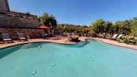This amenity area offers two distinct spaces designed for recreation and fitness. The outdoor pool area showcases a freeform design with crystal-clear turquoise water, complemented by a natural stone water feature at its center. The surrounding deck provides ample space for lounging, furnished with chaise lounges and shaded by vibrant red market umbrellas. Mature trees and lush landscaping frame the area, while a wrought iron fence encloses the space, offering privacy and security. Stone architectural elements in the background add a sophisticated, resort-like aesthetic under open Texas skies.

Adjacent to the pool, the fitness center is a bright, well-organized space featuring bold orange and blue accent stripes along the walls, giving it an energetic atmosphere. Large windows overlook the pool area, filling the room with natural light. The gym is comprehensively equipped with multiple treadmills, elliptical trainers, stationary and recumbent bikes, a multi-station strength training machine, and a full rack of dumbbells. A large wall mirror enhances the sense of space and allows for form monitoring during workouts. Durable carpet tile flooring and ceiling fans contribute to a comfortable exercise environment.