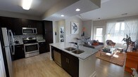 This single-floor residence offers a thoughtfully designed open-concept layout that seamlessly connects the kitchen, dining, and living areas into one cohesive, flowing space. The kitchen serves as the centerpiece, anchored by a large island with a double stainless steel sink and elegant granite countertops in a warm grey tone. Dark espresso cabinetry provides striking contrast against the light-colored walls, while stainless steel appliances — including a range, microwave, and dishwasher — add a sleek, modern touch. A mosaic tile backsplash adds a layer of texture and visual interest to the cooking area. Light hardwood flooring extends throughout the main living space, enhancing the open and airy feel. The dining area sits adjacent to the living room, which benefits from natural light streaming in through a large exterior door or window. The living area is comfortably proportioned and features a warm, inviting ambiance. Recessed lighting and track lighting illuminate the space evenly, complementing the natural light. A hallway visible from the main area hints at additional private rooms beyond, completing this well-appointed and functional single-level home.
