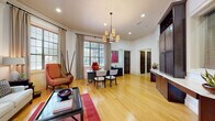 This single-floor professional space is designed to offer a polished and functional environment for both staff and visitors. The entry lounge sets a refined tone with warm hardwood flooring, tall windows dressed in linen drapes, and an arched transom above elegant glass-paneled doors that invite abundant natural light. A comfortable white sofa, upholstered armchairs, and a glass-top coffee table create a welcoming reception area with a refined residential feel.

Adjacent to the lounge, a spacious open area serves as a central hub, illuminated by a multi-shade pendant chandelier and recessed lighting. Crown molding and high ceilings add architectural character throughout. A dedicated private office features a sleek dark executive desk positioned beneath a large window, a lateral filing cabinet, and vibrant wall art, providing a focused and professional workspace.

A well-equipped conference and workroom includes a round table surrounded by bold red leather chairs, a built-in desk with dark cabinetry and upper storage, and generous window views of the surrounding greenery. Dark wood built-in cabinetry along one wall provides ample storage and countertop workspace. The consistent use of hardwood floors, neutral wall tones, and rich wood accents ties the entire space together in a cohesive, professional aesthetic.