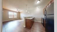 This efficiently designed 2-bedroom, 1-bathroom residence is laid out across a single floor, offering a practical and cohesive living experience. The open-concept kitchen and living area forms the heart of the home, anchored by a peninsula with granite-look countertops, rich dark wood cabinetry, pendant lighting, and black appliances including a refrigerator, range, and over-the-range microwave. Durable hardwood-style flooring extends throughout the main living areas, complemented by a ceiling fan that adds comfort to the living room, which receives natural light through a window fitted with blinds. A hallway with patterned carpet and a wall sconce connects the main living area to the private rooms. Both bedrooms feature plush carpeting and large windows that allow generous natural light to fill each room. The primary bedroom includes a ceiling fan and a closet accessible via a paneled white door. The shared bathroom is well-appointed with wood-look flooring, a full tub-shower combination with floor-to-ceiling earthy brown tile surround, a vanity with storage cabinetry above, and classic chrome fixtures. Neutral wall tones throughout create a clean, cohesive aesthetic that lends itself well to personalization.