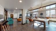 Located at 15 Poplar St in Glassboro, NJ, this contemporary single-floor community lounge is designed to foster both social interaction and productive work. The space boasts soaring ceilings that lend an open, airy atmosphere, complemented by large floor-to-ceiling windows that flood the interior with natural light and offer views of the surrounding exterior.

The main lounge area is anchored by rich cognac leather sofas and tufted seating arranged around a sleek glass coffee table atop a bold marbled area rug. Statement pendant chandeliers — including a dramatic circular halo fixture — serve as focal points overhead. A wall-mounted flat-screen TV and a textured brick accent wall add visual depth to the seating zone.

Adjacent dining areas feature round white tables paired with modern Scandinavian-style chairs, ideal for casual gatherings. A pool table provides additional recreational appeal. Throughout, wood-look tile flooring in a parquet-inspired pattern grounds the space with warmth and continuity.

A dedicated workstation nook, lined with geometric patterned wallcovering and equipped with bar-height desks and a mounted monitor, offers a functional retreat. Vibrant teal accent chairs and mosaic side tables introduce bold pops of color, creating an energetic yet inviting ambiance throughout this well-appointed communal space.