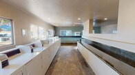 This single-floor communal laundry facility is designed for efficiency and convenience, offering a spacious, well-organized environment for residents. The room is lined along one wall with a continuous row of white top-load washing machines and dryers, providing generous laundry capacity. Opposite the machines runs an extended dark granite-topped folding counter, offering ample surface space for sorting and folding laundry. The flooring throughout is a durable, earth-toned tile that complements the clean, functional aesthetic of the space. Natural light pours in through multiple large windows positioned along the exterior walls, brightening the interior and creating an open, airy atmosphere. Recessed ceiling lights supplement the natural light, ensuring the space is well-lit at all times. The textured ceiling and soft sage-green accent walls add a subtle touch of warmth to the otherwise utilitarian setting. A wall-mounted heating or cooling unit is visible near the windows, ensuring year-round comfort. The layout is thoughtfully designed with a wide central corridor, allowing for easy movement and simultaneous use by multiple occupants. Overall, this laundry facility is practical, clean, and comfortable.