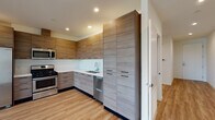 This contemporary single-floor residence showcases a clean, modern design with light wood-tone flooring carried consistently throughout the space, creating a cohesive and inviting atmosphere. The kitchen is a standout feature, designed in an L-shaped layout with floor-to-ceiling cabinetry finished in a warm wood-grain laminate. A full suite of stainless steel appliances includes a gas range, over-the-range microwave, dishwasher, and a side-by-side refrigerator with an ice and water dispenser. A white countertop with an undermount sink and recessed lighting complete the kitchen's polished, functional aesthetic. The living and bedroom areas are bright and open, featuring neutral walls, recessed lighting, and windows that bring in natural light. One room offers direct outdoor access through a sliding glass door that opens to a balcony with horizontal railing and views of mature trees. The residence includes two bathrooms: one with a single-sink vanity featuring wood-tone cabinetry and a tub-shower combo with a glass sliding door, and a second featuring a dual-sink vanity with a white countertop and a separate walk-in glass-enclosed shower. Both bathrooms maintain the same modern material palette seen throughout the home.