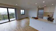 This contemporary single-floor space offers a clean, modern aesthetic defined by an open-concept design that connects the kitchen and living areas effortlessly. Light hardwood flooring flows throughout, enhancing the sense of openness and warmth across the entire layout. Recessed lighting is thoughtfully placed in the smooth white ceilings, providing consistent illumination throughout the space.

The kitchen features a sleek, stainless steel refrigerator and a white peninsula countertop that doubles as a casual bar or prep area, creating a natural boundary between the kitchen and the living space without compromising the open feel. Neutral white walls keep the interior bright and versatile, lending itself well to a variety of furnishing styles.

Natural light is a standout feature, streaming in through multiple windows and a large sliding glass door that leads to a private balcony. The balcony access frames panoramic outdoor views of the surrounding treetops and landscape, bringing a strong connection to the outdoors into the heart of the living area. The overall ambiance is fresh, airy, and functional — well-suited for those seeking a modern, low-maintenance living environment with an emphasis on light, openness, and clean design.