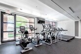 This expansive fitness center is designed to support a full range of workout routines across a thoughtfully organized, single-floor layout. The facility is divided into distinct training zones, each tailored to a specific type of exercise, creating a structured and efficient environment for users of all fitness levels.

The cardio area is positioned near large floor-to-ceiling windows that flood the space with natural light and offer views of lush greenery outside. This zone is equipped with a row of treadmills, elliptical trainers, and upright and recumbent stationary bikes — including Matrix-brand machines — complemented by wall-mounted flat-screen televisions for entertainment during workouts.

Moving through the facility, a dedicated strength training corridor features a comprehensive selection of selectorized machines, including Technogym equipment for targeted muscle group training. Structural columns punctuate the space and are integrated seamlessly into the layout. Floor-to-ceiling mirrors line portions of the walls, enhancing the sense of depth and allowing for form monitoring during lifts.

A free weights area includes a full dumbbell rack and adjustable benches, while a functional training zone offers stability balls, medicine balls, a squat/power rack, and open floor space for dynamic movement. Rubber flooring throughout provides safety and durability. A beverage vending machine adds convenience. The space is finished with a neutral grey palette, recessed and panel lighting, and white trim detailing.