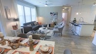 This single-floor residence showcases an open-concept design that seamlessly connects the dining area, living room, and beyond. The dining space, positioned at the foreground, features a well-set table dressed with warm orange placemats, coordinating dishware, and decorative centerpieces, creating an inviting atmosphere for entertaining. The living area flows naturally from the dining space and is anchored by a tufted dark gray sectional sofa, a round white coffee table, and a patterned area rug. Two gray accent chairs complete the seating arrangement, adding a balanced, curated feel. Wood-look flooring extends throughout the space, providing a cohesive visual foundation. A ceiling fan overhead ensures comfort while contributing to the room's clean, contemporary aesthetic. Large windows dressed with white blinds allow natural light to fill the space, enhancing the neutral gray and beige color palette. A hallway visible in the background leads to additional rooms, and a bar-height counter with stools is partially visible to the right, suggesting a kitchen nearby. The overall ambiance is modern, warm, and thoughtfully designed.
