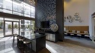This single-floor space makes a striking impression through its dramatic double-height ceilings, which draw the eye upward and lend an expansive, airy quality to the interior. The focal point of the room is a floor-to-ceiling textured brick feature wall rendered in deep charcoal tones, providing a bold architectural backdrop for a mounted flat-screen television and a sculptural metallic centerpiece that adds artistic flair.

A sleek kitchen or bar area is positioned prominently, featuring dark granite countertops, a built-in beverage refrigerator with stainless steel cabinetry below, and bar-height seating with modern stools — ideal for casual gathering or entertainment. Large floor-to-ceiling windows flood the space with abundant natural light while framing views of the exterior courtyard and surrounding buildings, seamlessly connecting the indoors with the outside environment.

On the opposite side, a comfortable lounge seating area with mustard-toned upholstered benches and small side tables creates a relaxed social zone. Decorative metallic wall accents and a contemporary chandelier overhead contribute to the polished, modern aesthetic. Light-toned tile flooring grounds the space with a clean, refined finish throughout.