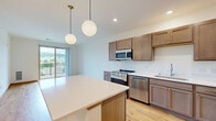 This well-appointed 2-bedroom, 1-bathroom unit in Middleton, Wisconsin offers a thoughtfully designed single-floor layout filled with natural light and modern finishes throughout. The open-concept kitchen and living area serves as the heart of the home, featuring warm-toned hardwood-style flooring, taupe shaker cabinetry, stainless steel appliances including a gas range, dishwasher, microwave, and refrigerator, and a central island with a crisp white countertop accented by stylish globe pendant lighting. The living area flows seamlessly toward a sliding glass door that opens onto a private balcony, offering elevated views of the surrounding Middleton neighborhood and lush greenery. Both bedrooms are carpeted for comfort and feature double-door closets with warm oak wood trim that ties into the overall design palette. Large windows with blinds allow for ample natural light while framing pleasant outdoor views. The bathroom is clean and functional, offering warm wood cabinetry, a white countertop vanity with gold-toned hardware, light tile flooring, and black accent fixtures. Crisp white walls and warm wood trim details create a cohesive, modern aesthetic throughout this comfortable and efficiently designed home.