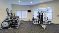 This fitness center is designed to accommodate a variety of workout routines within a single, open floor layout. The room is finished with neutral light-colored walls and durable patterned carpet flooring suited for high-traffic use. Recessed lighting provides consistent overhead illumination throughout the space, complemented by natural light streaming in through multiple windows fitted with white horizontal blinds.

The cardio section features several elliptical machines and a treadmill, offering options for sustained aerobic exercise. A multi-station strength training machine occupies the opposite side of the room, enabling a range of resistance-based exercises. A weight bench and floor mat provide additional space for targeted workouts and stretching. A portable fan is available for added air circulation during intense sessions.

A wall-mounted flat-screen television is positioned at an accessible viewing angle, ideal for entertainment or instructional content during workouts. A wall clock keeps users aware of time. The overall layout is organized and spacious, with clear pathways between equipment ensuring ease of movement and safety. The combination of natural light, functional equipment, and a clean, practical design makes this fitness center a well-rounded facility for everyday exercise needs.