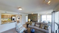 This single-floor residence offers an open-concept layout that connects the main living areas in a cohesive and functional design. The living room serves as the central hub, featuring neutral-toned carpeted flooring and a notably accented dark feature wall that adds depth and visual contrast to the space. Flush-mount and ceiling fan light fixtures provide ambient illumination throughout.

The living area comfortably accommodates a full seating arrangement, with a view into the dining area near a bright window dressed with grommet-panel curtains, allowing natural light to filter in. A doorway from the living room leads to additional rooms beyond, suggesting a practical and well-organized floor plan.

The kitchen is visible from the living space through an open pass-through area, showcasing white cabinetry, a dark countertop, and full-sized appliances including a refrigerator with an integrated ice and water dispenser. A small dining nook with a table and chairs is nestled adjacent to the kitchen.

Overall, the space presents a warm, welcoming ambiance with a balance of natural light, modern fixtures, and a neutral color palette accented by thoughtful design details.
