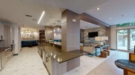 This single-floor amenity space is designed with a contemporary aesthetic that balances functionality and refined style throughout its interconnected zones. Signature geometric-patterned porcelain tile flooring unifies the entire space, complemented by warm wood-paneled columns and cabinetry that provide visual continuity from one area to the next.

The fully equipped kitchen runs along one side of the space and features dark stone countertops, stainless steel appliances including a full-size refrigerator and dishwasher, an undermount sink, and ample cabinetry with open shelving. Linear LED lighting overhead ensures the workspace is well-lit and functional.

Adjacent to the kitchen, a lounge area offers tufted teal banquette seating set against a stacked-stone accent wall, paired with dark marble-topped tables — an ideal setting for casual gatherings. A dedicated sitting area nearby showcases a striking hand-painted mural accent wall depicting a mountainous landscape, anchored by neutral-toned sofas, a live-edge wood coffee table, and a wall-mounted television.

The billiards room features a full-size pool table beneath a sleek pendant light, a mounted television, and stone accent walls. Floor-to-ceiling glass doors throughout flood the space with natural light and offer views of lush outdoor greenery.