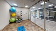 This single-floor fitness studio is thoughtfully designed to support a wide range of exercise and rehabilitation activities. The space is defined by floor-to-ceiling aluminum-framed glass walls and doors, creating an open, transparent feel that connects the studio visually to an adjacent main gym area equipped with cardio machines and additional fitness equipment. This architectural detail promotes a sense of openness while maintaining distinct, functional zones.

The studio's interior features durable wood-look plank flooring, which provides both aesthetic warmth and a practical surface for floor-based exercises. A drop ceiling with recessed panel lighting ensures consistent, even illumination throughout the space. One wall is equipped with a large wall-mounted flat-screen television along with a smaller secondary monitor, supporting guided workouts or instructional content.

A dedicated equipment storage area houses an organized multi-tier rack stocked with medicine balls, resistance bands, stability balls in multiple sizes, foam rollers, and exercise mats. This comprehensive selection of functional training tools makes the space well-suited for personal training sessions, group fitness classes, or physical therapy. The overall ambiance is clean, modern, and professional, reflecting a purposeful design focused on fitness functionality.