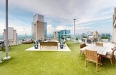 The SHN Rooftop Outdoor Lounge is a 680-square-foot open-air amenity space situated atop a high-rise building, offering sweeping panoramic views of the surrounding cityscape and skyline. The rooftop is finished with lush artificial turf that creates a vibrant, park-like atmosphere elevated above the urban landscape. A central seating area surrounds a stone-clad fire pit, complemented by stylish outdoor sofas, accent chairs with blue cushions, and rounded wicker lounge pods ideal for relaxed gatherings. An outdoor dining area features a wooden table with wicker chairs positioned beneath a wall-mounted flat-screen television, accented by a light stone facade that adds architectural texture. A built-in grill station with additional seating is available for outdoor cooking and entertaining. Transitioning indoors, a bright enclosed lounge corridor features large floor-to-ceiling glass walls and doors, a wall-mounted TV above a built-in fireplace, and large-format tile flooring, providing a climate-controlled retreat with continued city views. The entry vestibule offers a clean, minimalist aesthetic with smooth white walls and tiled flooring. The overall ambiance of the space is modern, sophisticated, and resort-inspired, designed for social gatherings and relaxation at elevation.