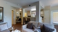 This single-floor residence offers a thoughtfully designed open-concept layout that seamlessly connects the main living spaces. The living area serves as the heart of the home, featuring rich hardwood-style flooring, neutral-toned walls, and comfortable seating accented with decorative pillows that add a touch of modern style.

A raised breakfast bar with dark wooden bar stools separates the living area from the kitchen, which is equipped with white cabinetry, a full-size refrigerator, and a built-in microwave. The kitchen's warm lighting creates an inviting atmosphere well-suited for both cooking and casual dining.

Tucked just off the main living space is a dedicated home office nook, complete with a desk and ergonomic office chair, making it a practical and private area for work or study. Framed artwork and a small plant add personality and warmth to this corner.

The space benefits from recessed and overhead lighting throughout, ensuring each area is well-lit. The consistent use of warm neutral tones, dark wood flooring, and clean architectural lines gives the residence a cohesive and contemporary aesthetic. The open flow between spaces makes the layout feel expansive and functional, ideal for everyday living.