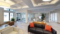 This well-appointed single-floor space functions as a sophisticated communal lounge and gathering area, thoughtfully designed to balance both comfort and functionality. The main lobby area immediately commands attention with its striking coffered ceiling, which incorporates recessed lighting and an elegant oversized drum pendant light. Tall, floor-to-ceiling windows along the front facade flood the interior with abundant natural light while offering views of the surrounding streetscape. Clerestory windows positioned above the primary window line further enhance the brightness throughout.

The seating areas are arranged into distinct zones, featuring a combination of cream-toned upholstered sofas and armchairs alongside woven accent chairs grouped around a circular white coffee table. A wall-mounted flat-screen television, a wooden media console, and bold original artwork — including a vibrant abstract canvas and a yellow statement piece — contribute to the curated, gallery-like atmosphere.

A tiled gas fireplace serves as a warm focal point in one section of the space, topped by a large-scale colorful abstract painting. Adjacent to the lobby, a glass-walled meeting room provides a semi-private workspace with a glimpse into an active professional environment.

Sliding glass doors at the rear of the space open to a furnished outdoor terrace, extending the usable area and inviting natural airflow. Light wood and tile flooring unify the entire space cohesively.