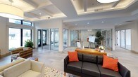 This well-appointed single-floor space functions as a sophisticated communal lounge and gathering area, thoughtfully designed to balance both comfort and functionality. The main lobby area immediately commands attention with its striking coffered ceiling, which incorporates recessed lighting and an elegant oversized drum pendant light. Tall, floor-to-ceiling windows along the front facade flood the interior with abundant natural light while offering views of the surrounding streetscape. Clerestory windows positioned above the primary window line further enhance the brightness throughout.

The seating areas are arranged into distinct zones, featuring a combination of cream-toned upholstered sofas and armchairs alongside woven accent chairs grouped around a circular white coffee table. A wall-mounted flat-screen television, a wooden media console, and bold original artwork — including a vibrant abstract canvas and a yellow statement piece — contribute to the curated, gallery-like atmosphere.

A tiled gas fireplace serves as a warm focal point in one section of the space, topped by a large-scale colorful abstract painting. Adjacent to the lobby, a glass-walled meeting room provides a semi-private workspace with a glimpse into an active professional environment.

Sliding glass doors at the rear of the space open to a furnished outdoor terrace, extending the usable area and inviting natural airflow. Light wood and tile flooring unify the entire space cohesively.