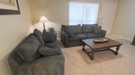 This single-floor space centers around a well-appointed living area designed for comfort and functionality. The room features two large, plush dark grey sofas arranged to encourage conversation, complemented by a classic wooden coffee table with turned legs that serves as a sturdy focal point. Neutral beige carpeting runs throughout, contributing to the warm, understated ambiance of the space. A floor lamp with a warm-toned shade is positioned in the corner alongside a small side table adorned with a modest houseplant, adding a touch of greenery to the room. A large window fitted with horizontal blinds allows natural light to filter in, brightening the interior while maintaining privacy. The walls are finished in a soft, muted tone that ties the furnishings together cohesively. The overall layout is open and uncluttered, making efficient use of the available floor space. The single-level design ensures easy accessibility throughout, and the thoughtful arrangement of furniture creates a relaxed, livable atmosphere suited for everyday use.