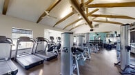 This fitness center offers a comprehensive workout environment designed to support a wide range of exercise routines. The space is immediately distinguished by its soaring vaulted ceilings adorned with exposed wooden beams and industrial-style ductwork, creating an open, airy atmosphere enhanced by ceiling fans throughout. Durable rubber flooring spans the entire facility, providing a safe and practical surface for all activities.

The cardio zone features multiple treadmills and elliptical machines, each positioned near large windows that allow ample natural light and outdoor views, creating an energizing environment for aerobic workouts. A wall-mounted flat-screen television is positioned for easy viewing during cardio sessions.

The strength training area is outfitted with a full lineup of Cybex VR1 selectorized machines, covering major muscle groups for a complete resistance training program. A dedicated free weights section includes a well-stocked dumbbell rack with a large wall mirror, ideal for monitoring form during lifts. Cable machines and a functional training station round out the strength offerings.

Full-length mirrors are strategically placed along the walls, amplifying the sense of space and supporting proper exercise technique. The light teal accent walls contribute a fresh, motivating aesthetic throughout the facility, making this fitness center a functional and inviting space for daily training.