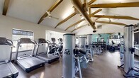 This fitness center offers a comprehensive workout environment designed to support a wide range of exercise routines. The space is immediately distinguished by its soaring vaulted ceilings adorned with exposed wooden beams and industrial-style ductwork, creating an open, airy atmosphere enhanced by ceiling fans throughout. Durable rubber flooring spans the entire facility, providing a safe and practical surface for all activities.

The cardio zone features multiple treadmills and elliptical machines, each positioned near large windows that allow ample natural light and outdoor views, creating an energizing environment for aerobic workouts. A wall-mounted flat-screen television is positioned for easy viewing during cardio sessions.

The strength training area is outfitted with a full lineup of Cybex VR1 selectorized machines, covering major muscle groups for a complete resistance training program. A dedicated free weights section includes a well-stocked dumbbell rack with a large wall mirror, ideal for monitoring form during lifts. Cable machines and a functional training station round out the strength offerings.

Full-length mirrors are strategically placed along the walls, amplifying the sense of space and supporting proper exercise technique. The light teal accent walls contribute a fresh, motivating aesthetic throughout the facility, making this fitness center a functional and inviting space for daily training.