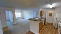This single-floor space is designed with an open-concept layout that connects the kitchen and living area into one cohesive, airy environment. The kitchen features a peninsula with a dark laminate countertop, a double-basin sink, warm wood-toned cabinetry, and white appliances, including a dishwasher and refrigerator. Hardwood-style flooring anchors the kitchen zone, adding warmth and contrast to the surrounding space. The living area transitions smoothly from the kitchen via a subtle flooring change to plush carpeting, creating a comfortable and defined zone for relaxation. A ceiling fan with a light fixture is centrally positioned in the living area, promoting air circulation throughout. A large window dressed with vertical blinds allows natural light to fill the space, contributing to a bright and welcoming atmosphere. White walls throughout maintain a clean, neutral aesthetic, while visible closet doors near the back of the living area suggest additional storage options. The overall layout is practical and well-proportioned, offering a functional flow between the kitchen and main living space.