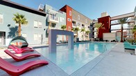 This outdoor amenity area is designed around a generously sized resort-style pool that serves as the centerpiece of the space. The pool features crystal-clear turquoise water and a striking architectural arch element rising from within, adding a sculptural focal point. Vivid pink lounge chairs line the pool deck, complementing the bold red, teal, and gray exterior facades of the surrounding multi-story buildings, which are accented by tall, mature palm trees that reinforce the resort aesthetic.

A dedicated spa area is positioned adjacent to the main pool, featuring a wall-mounted waterfall fixture that cascades into a tiled soaking basin, creating a tranquil retreat within the larger space. The pool deck is finished with clean, light-toned concrete pavers and is lined with teal-cushioned chaise lounges on both sides.

A covered cabana lounge anchors one end of the pool area, featuring a retractable louvered pergola roof strung with bistro lighting. Inside, dark wicker sectional seating is dressed in vibrant teal, coral, and striped cushions surrounding mirrored-top coffee tables. An additional covered lounge area equipped with a ceiling fan, stone-clad fireplace wall, and open glass doors offers a semi-enclosed gathering space with direct views of the pool and courtyard beyond.