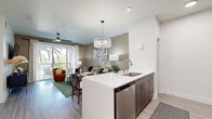 Located in Henderson, Nevada, this well-appointed one-bedroom, one-bathroom residence offers a thoughtfully designed open-concept floor plan with modern finishes throughout. Light wood-tone flooring flows seamlessly across the entire living space, complementing the clean white walls and creating a bright, cohesive aesthetic.

The kitchen serves as a central highlight, featuring a large island with a white quartz countertop, an undermount sink, stainless steel appliances including a refrigerator, microwave, and dishwasher, dark wood cabinetry, and a striking crystal chandelier overhead. The kitchen opens directly into the living area, which benefits from access to a private balcony with views of palm trees, enhancing the indoor-outdoor connection. A ceiling fan adds comfort to the living space.

The entry hallway is accented by an art display wall and leads to a dedicated closet, offering practical storage. The spacious primary bedroom accommodates a large bed comfortably, and features a ceiling fan, warm neutral tones, and natural light from a large window. The adjoining bathroom is finished with a wood-grain vanity, quartz countertop, backlit mirror, and a glass-enclosed shower, all rendered in a clean, contemporary palette.