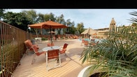 This outdoor amenity space centers around a generously sized swimming pool with clear, blue water and easy-access entry rails, making it both inviting and functional. The expansive pool deck is finished in a warm-toned concrete surface and is well-furnished with terracotta-hued lounge chairs and tables topped with large market umbrellas in coordinating shades of orange and tan, providing ample shade for relaxation. Mature palm trees and other established greenery are planted throughout the deck area, adding a resort-like atmosphere to the setting. The pool area is enclosed by a perimeter fence, ensuring privacy and security, while the surrounding multi-story buildings frame the space. Adjacent to the pool, a thoughtfully designed courtyard features modern vertical-bar metal fencing in a warm bronze tone, complemented by curated landscaping that includes ornamental grasses, broadleaf plants, and layered ground cover. Wooden accent walls add a natural, contemporary touch to the courtyard's design. The overall ambiance of this outdoor space is relaxed and sun-drenched, with a California-casual aesthetic that blends functional amenities with attractive landscaping for a comfortable and visually appealing environment.