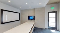 The conference room at 2510 West Ranch in Carlsbad is a professionally designed meeting space built to support productive collaboration. The room is anchored by a long, white stone-topped central table surrounded by multiple ergonomic chairs, offering generous seating capacity for teams and clients alike. The flooring features a rich, dark-toned carpet that contrasts elegantly with the room's bright, neutral palette. One wall is clad in large-format, warm-toned stone tiles, lending the space a refined, architectural character. Mounted at the center of this feature wall is a built-in flat-panel display, ideal for presentations and video conferencing. To the side, a large projection screen provides an additional viewing surface, making the room well-equipped for a variety of presentation formats. Recessed ceiling lighting ensures even, professional illumination throughout the space. The overall design strikes a balance between clean minimalism and polished sophistication, creating an environment that is both functional and visually impressive — well-suited for board meetings, client presentations, and collaborative sessions.