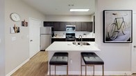 This thoughtfully designed 1-bedroom residence offers a cohesive blend of modern style and functional living across a single floor. The open-concept kitchen serves as a central hub, featuring dark wood cabinetry, subway tile backsplash, stainless steel appliances, and a large white quartz island with bar seating — ideal for casual dining or entertaining. Wood-look flooring flows through the kitchen and into the dining area, where large windows dressed with sheer white curtains and wood blinds invite natural light and views of surrounding greenery. A bold teal accent wall adds a vibrant design touch to the dining space. The living room continues the contemporary aesthetic with deep charcoal walls, a ceiling fan, hardwood-style flooring, and a generously sized window that fills the room with natural light. The bedroom is a calm retreat, appointed with soft carpeting, a ceiling fan, and ample natural light from a curtained window. A dedicated walk-in closet provides generous storage, while an additional door connects to a private bathroom. The bathroom features a dark vanity with a white quartz countertop, a framed mirror, and hardwood-style flooring, with convenient access to a well-organized linen closet.